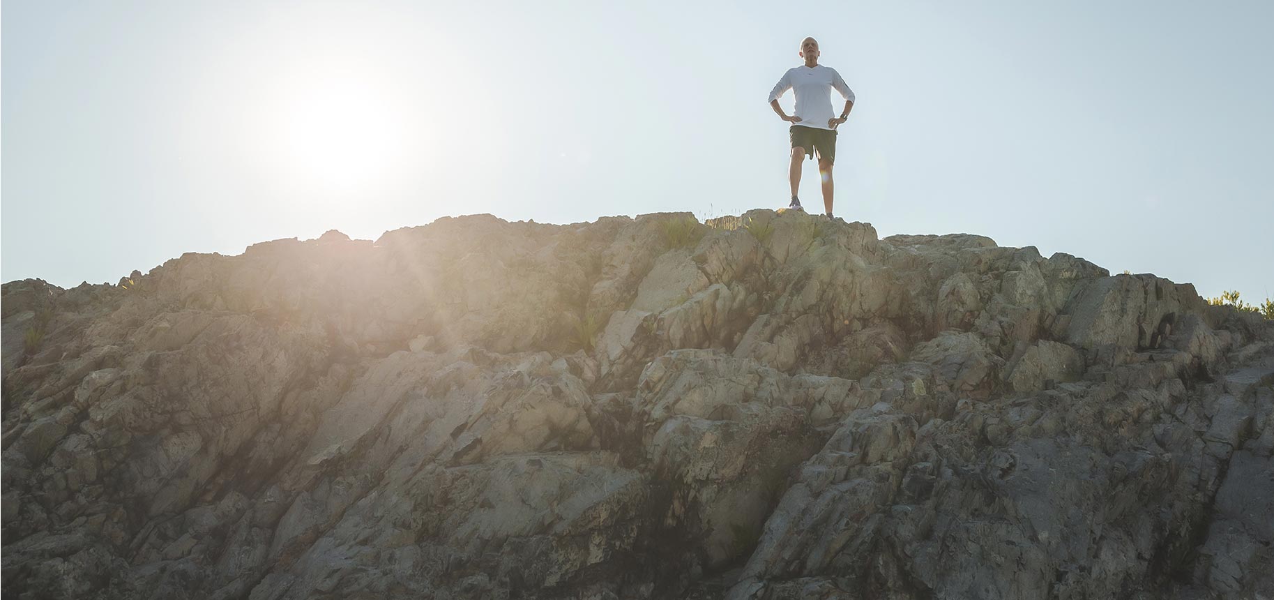 Marcy Schwam with hands on her hips atop a mountain with the sun shining behind.