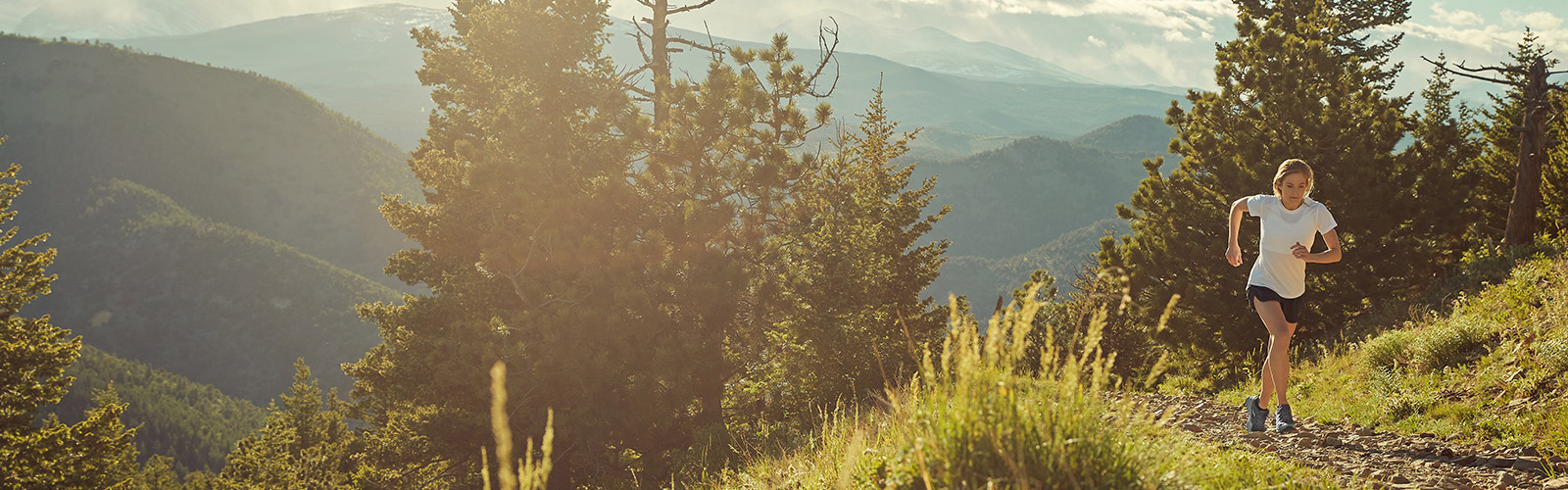 Runner hits a hairpin curve while coming down a dirt trail between trees on the side of a grassy mountain.