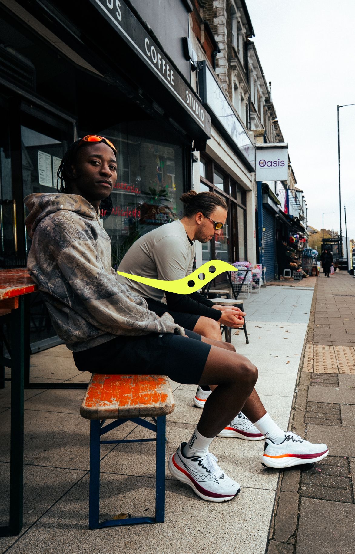 Two people in athletic wear sit on benches outside a shop on a city sidewalk. Both are wearing running shoes and sunglasses; one looks at the camera while the other looks at a phone.