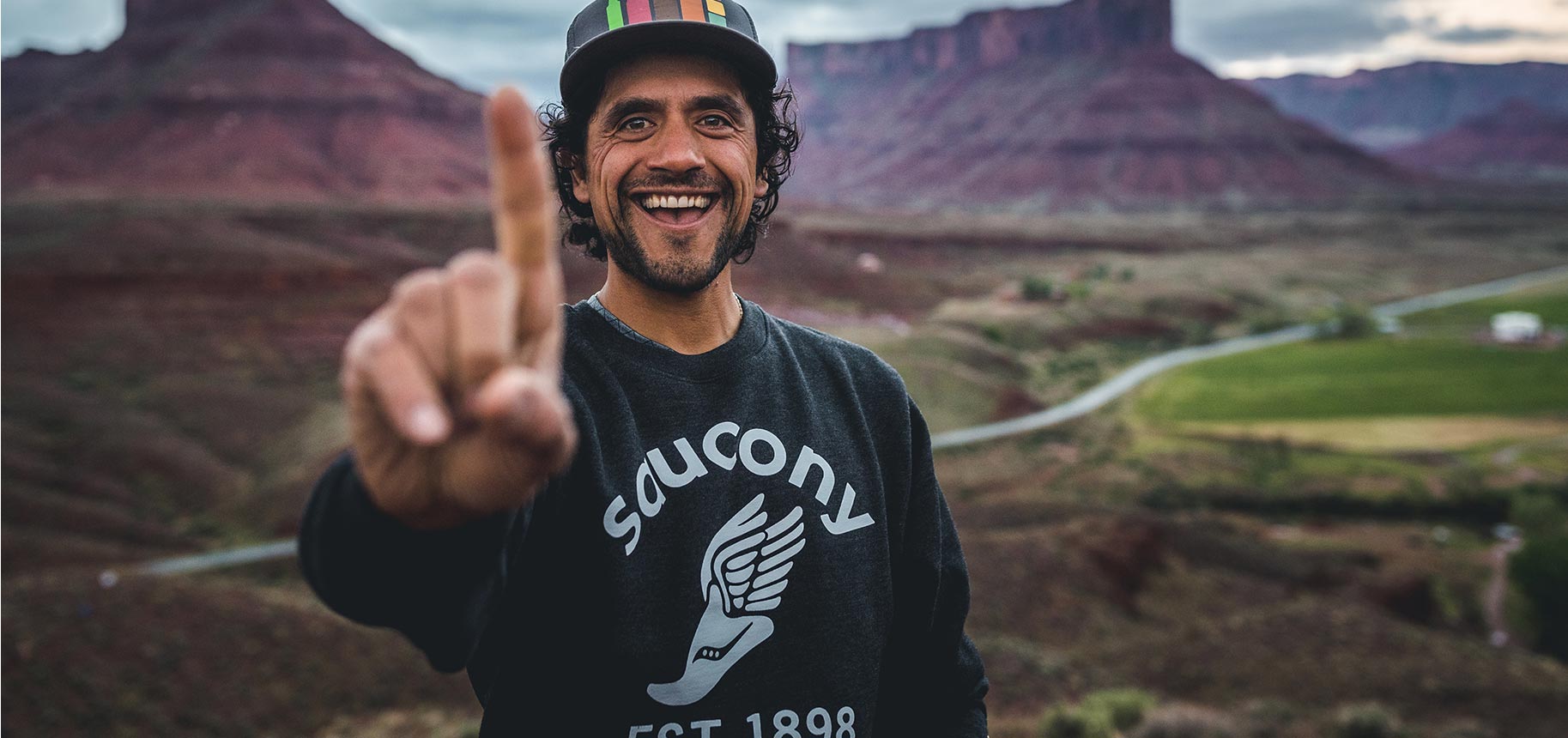 Eduardo Garcia's smiling face in a rainbow hat and Saucony EST 1898 sweatshirt showing his index finger as number 1.