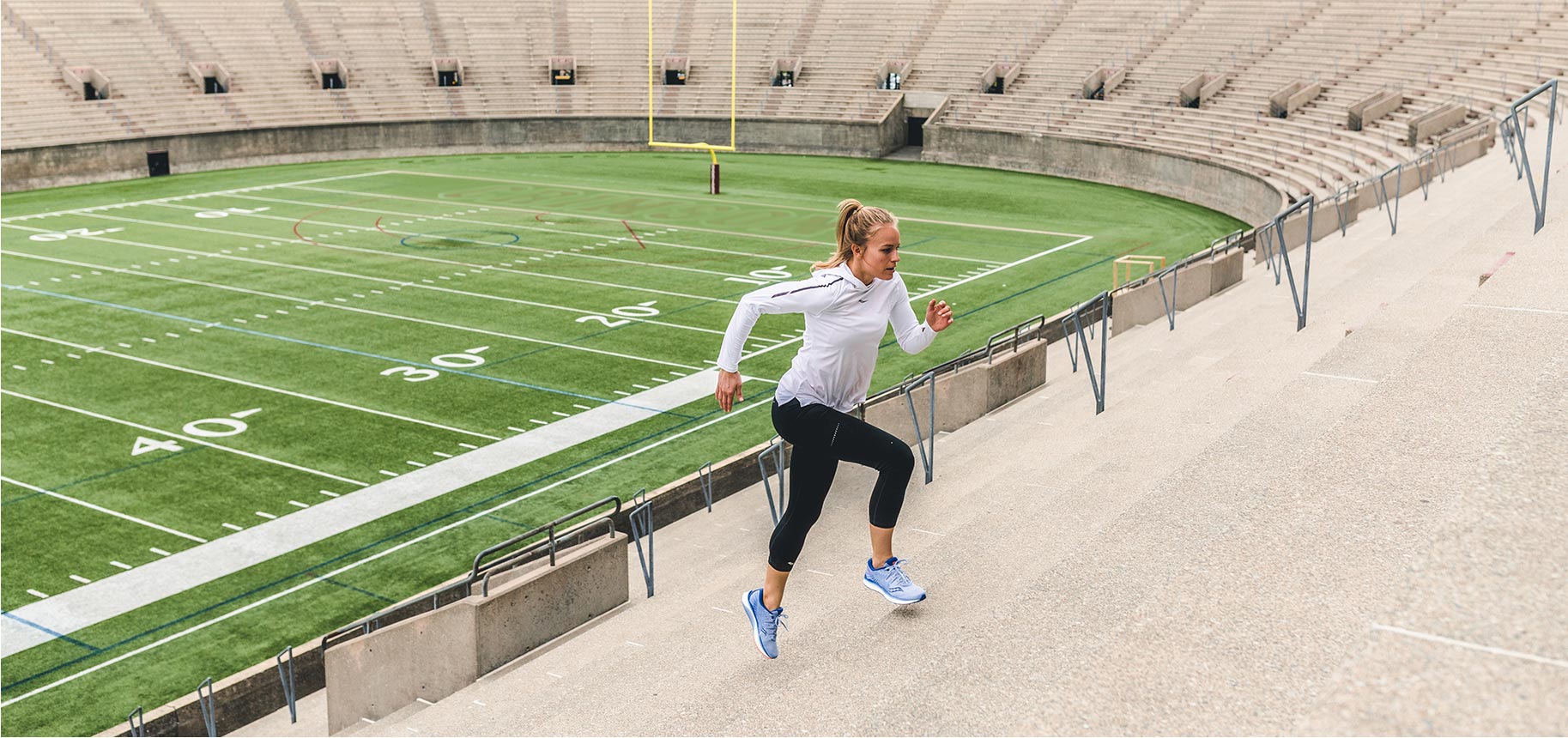 Daisy Robinton running in blue Saucony shoes up concrete stadium stairs with a football field in the background.