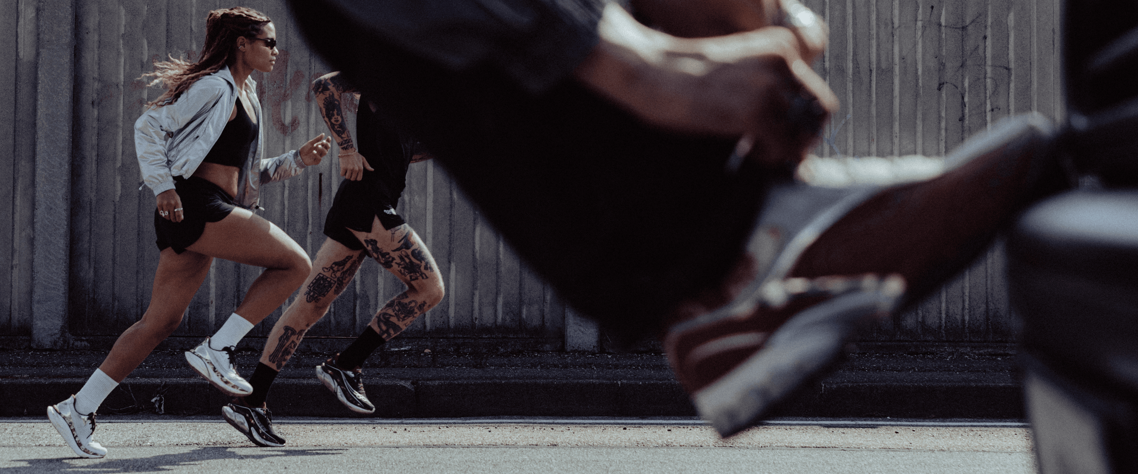 Two runners in Saucony Space Cowboy collection shoes and apparel running past a concrete wall.