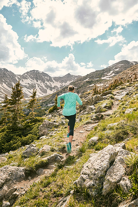 Woman running up a grassy mountainside. Blue skies above, a mountain range and pine trees visible in the distance.