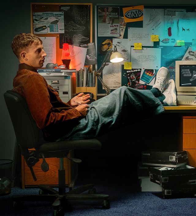 Man weating a brown jacket and wide jeans with his feet up on a desk beside an old computer.