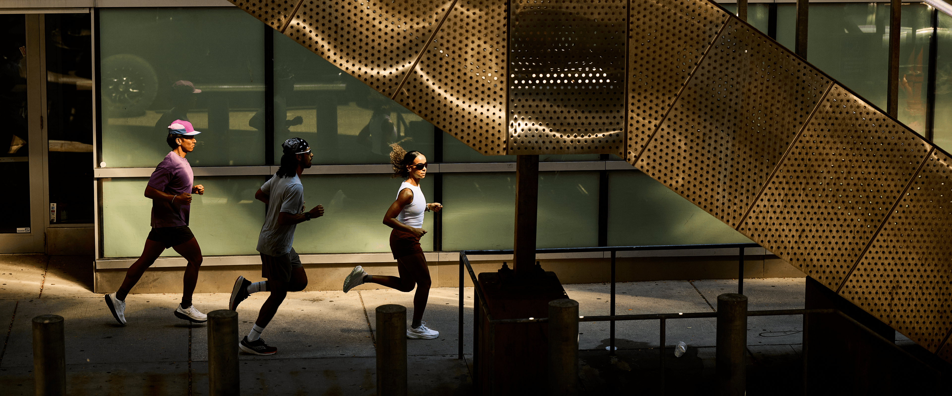 Three runners mid-stride on a city sidewalk past a glass building.