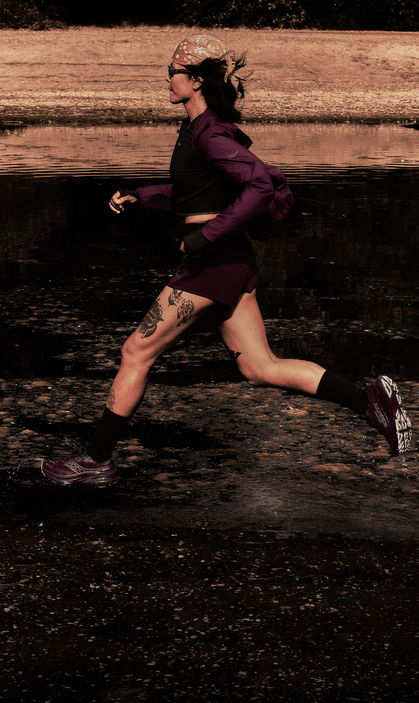 A runner in a maroon jacket and shorts splashing through a shallow stream at sunset.