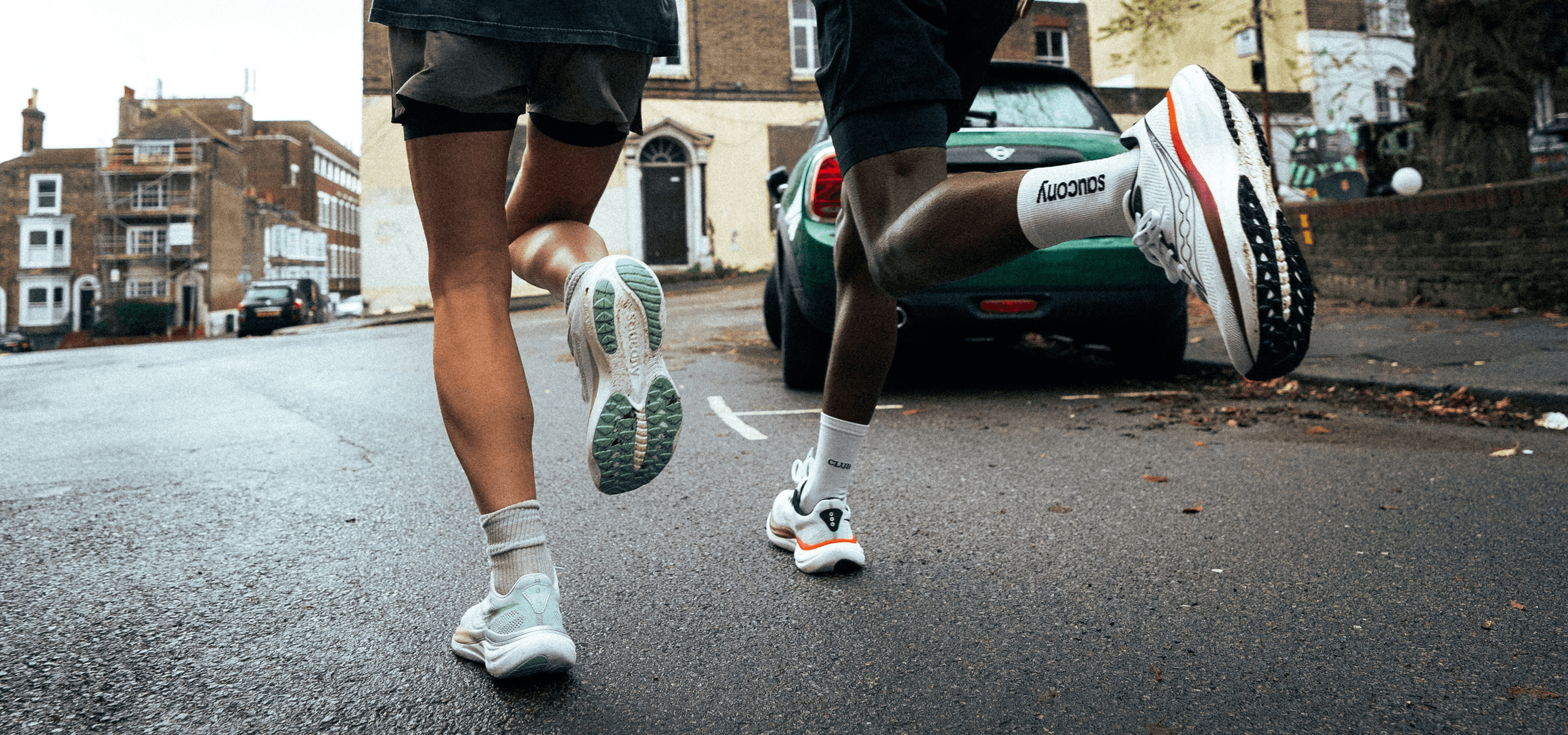 Two runners' legs and Saucony running shoes mid-stride on a wet city street.