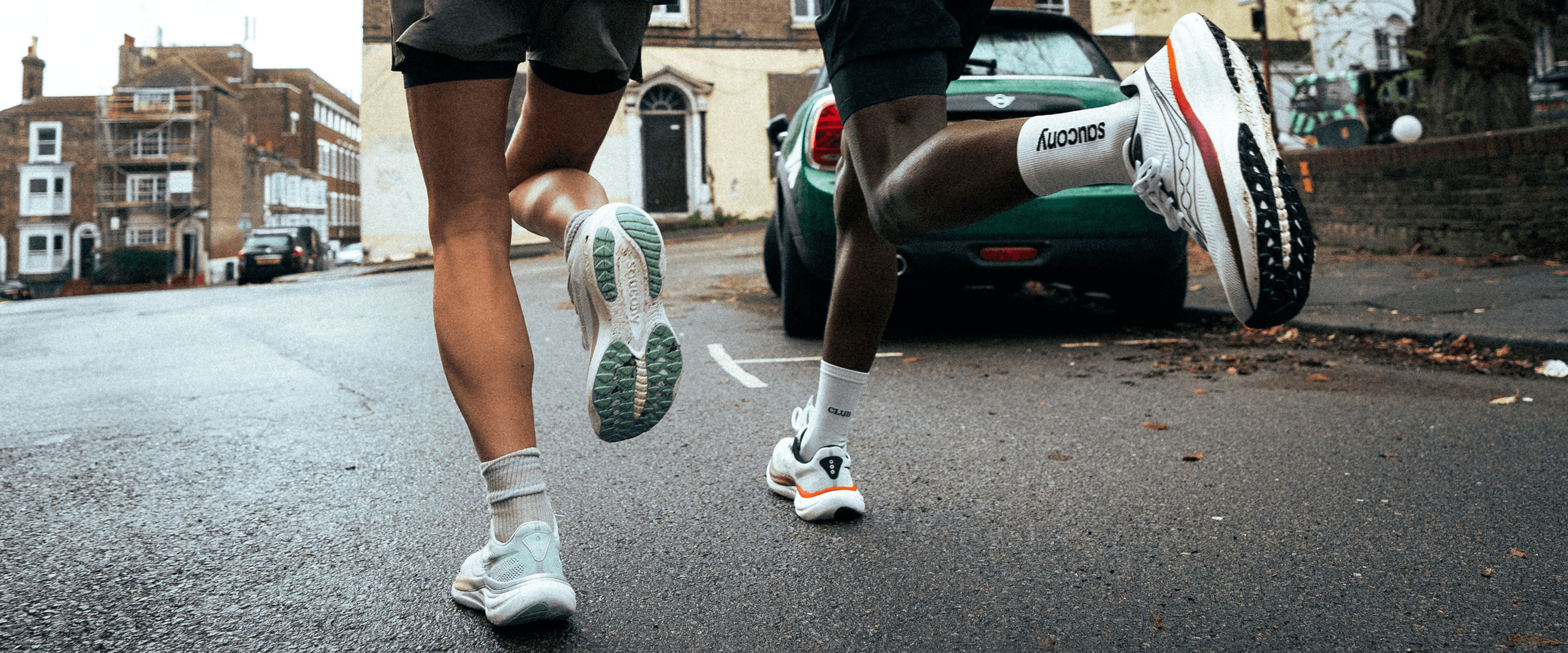 Two runners' legs and Saucony running shoes mid-stride on a wet city street.