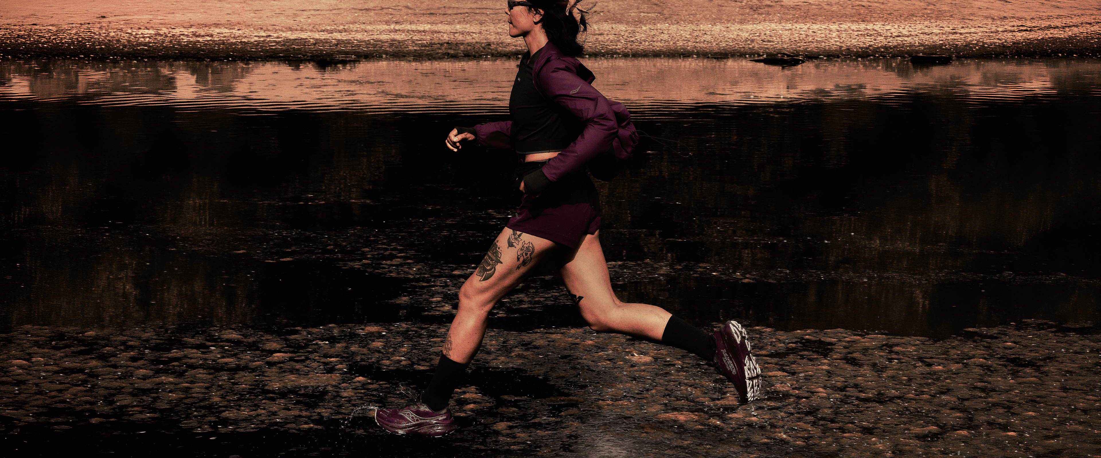 A runner in a maroon jacket and shorts splashing through a shallow stream at sunset.
