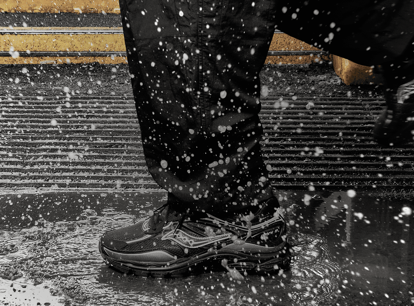 A close-up of black Saucony running shoes splashing through rain on a city street.